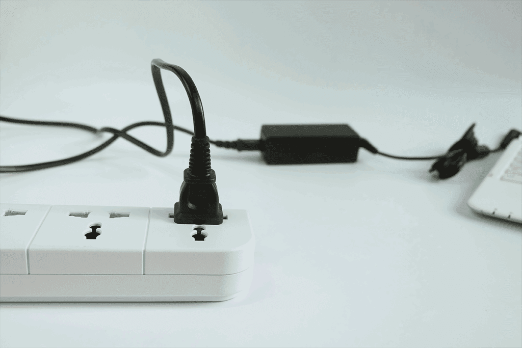 White power strip receives a black plug; cable snakes to a rectangular laptop power brick and a partially visible laptop on a clean white surface.